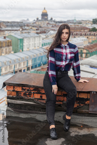 An Armenian girl with flowing long black hair in a checkered shirt sits on a rooftop in the center of St. Petersburg