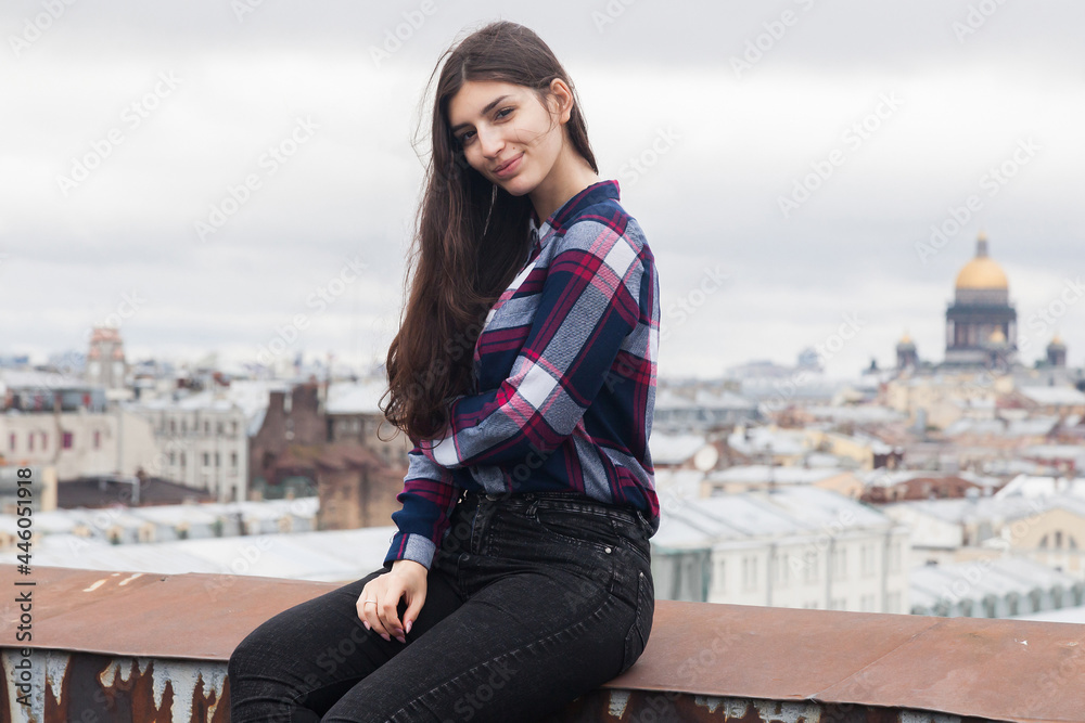 Fototapeta premium portrait of an Armenian girl with fluttering long black hair in a checkered shirt and jeans on a rooftop in the center of St. Petersburg