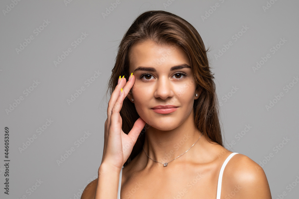 Portrait of beautiful brunette with bare shoulders touching her cheek, looking at camera and smiling, on gray background Portrait of beautiful brunette with bare shoulders touching her cheek, looking at camera and smiling, on gray background