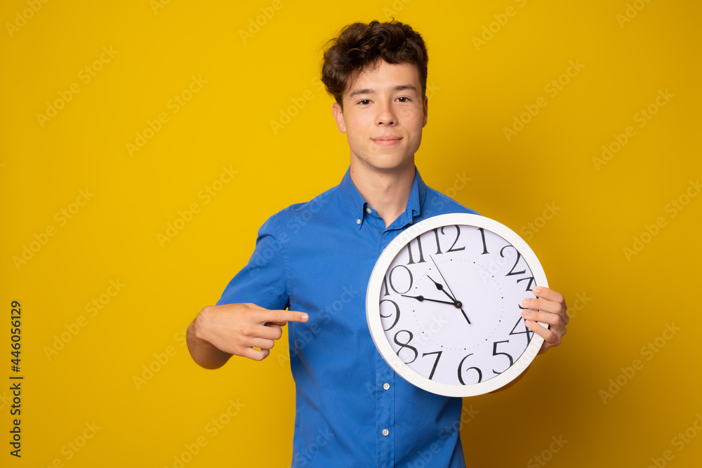 Portrait of caucasian teen boy with clock. Funny teenager showing clock, looking at camera. Child back to school, isolated on yellow. Education and time concept. Portrait of caucasian teen boy with clock. Funny teenager showing clock, looking at camera. Child back to school, isolated on yellow. Education and time concept.