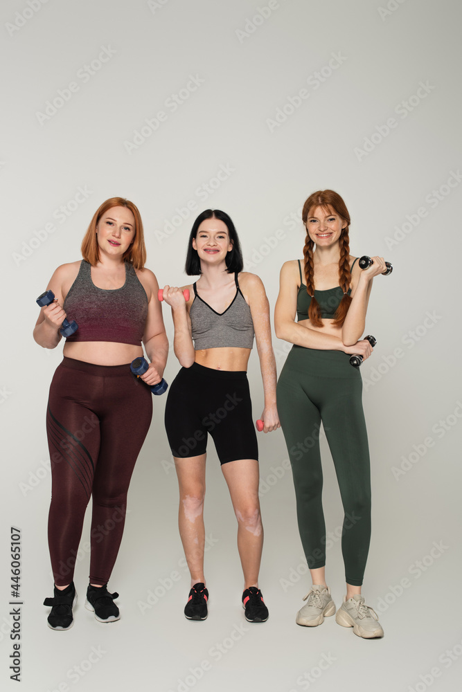 Fototapeta premium Smiling body positive sportswomen holding dumbbells on grey background