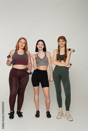 Smiling body positive sportswomen holding dumbbells on grey background