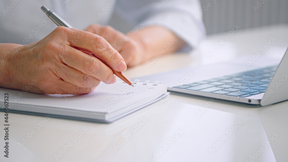 Old woman wrinkly hands write in paper notebook with pen at table near grey laptop keyboard Old woman wrinkly hands write in paper notebook with pen at table near grey laptop keyboard