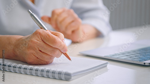 Senior lady teacher with wrinkly hands holds coloured pen over white page of paper notebook near grey laptop on table