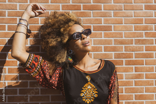 Beautiful African American woman wearing sunglasses leaning against  orange bricks wall tiding up her curly hair. Copy space for text. photo