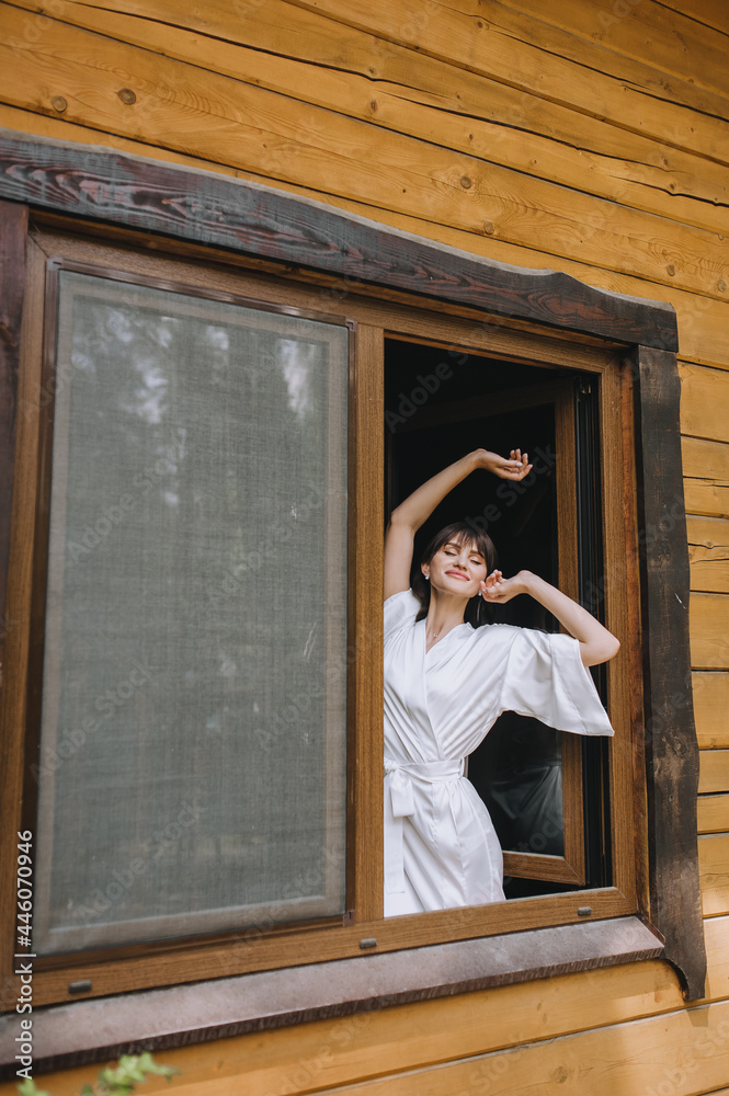 Fototapeta premium A beautiful, sleepy brunette bride in white lingerie with a long veil stands in the window against the background of the wall of a wooden house in the morning. Wedding photography.