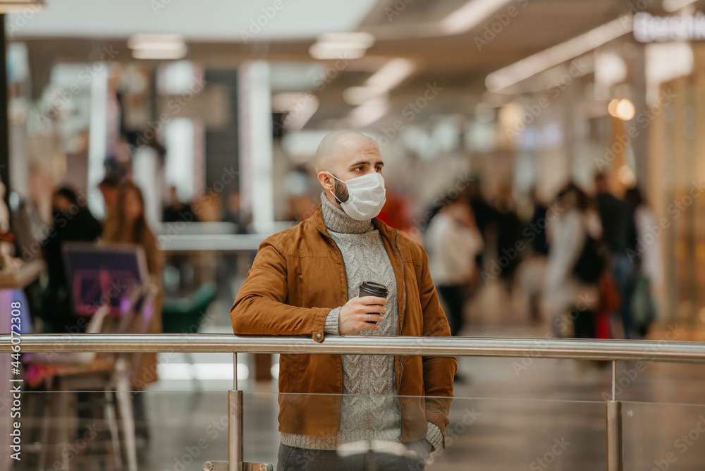 A man in a medical face mask to avoid the spread of coronavirus is holding a cup of coffee while waiting in the shopping center. A bald guy in a surgical mask is keeping social distance.