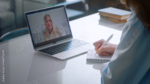 Medical university lady student in blue coat writes lecture in notebook sitting at online lesson via laptop with lecturer