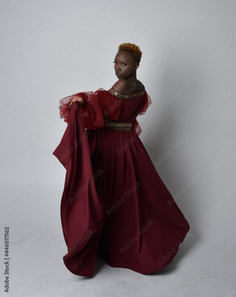 Full length portrait of pretty African woman wearing long red renaissance medieval fantasy gown, standing pose on a light grey studio background.