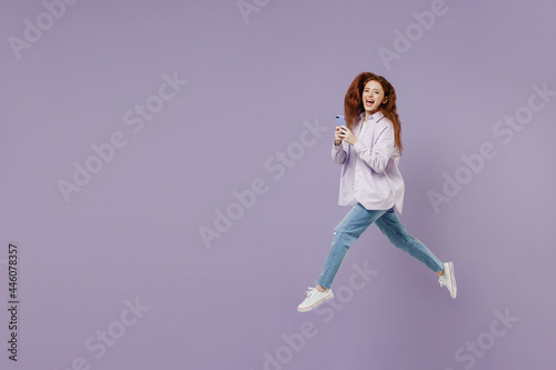 Full size body length laughing young redhead curly woman 20s wears white T-shirt violet jacket jump hold in hand use mobile cell phone isolated on pastel purple color wall background studio portrait.