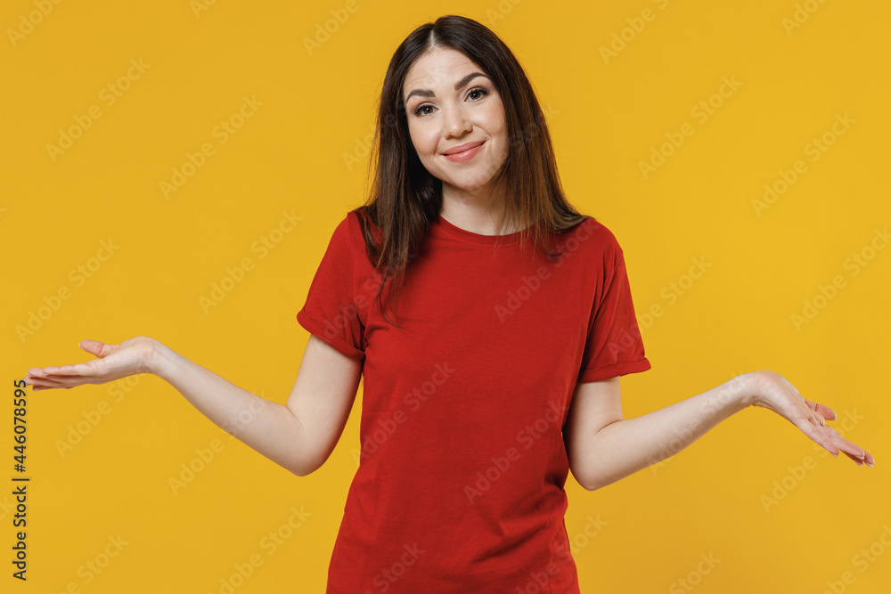 Smiling pensive puzzled confused beautiful young brunette woman 20s wears basic red t-shirt shrugging spreading hands isolated on yellow background studio portrait. People emotions lifestyle concept. Smiling pensive puzzled confused beautiful young brunette woman 20s wears basic red t-shirt shrugging spreading hands isolated on yellow background studio portrait. People emotions lifestyle concept.