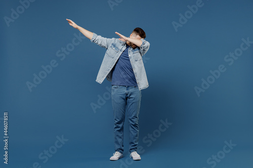 Full size length body happy handsome fun young brunet man 20s wears denim jacket doing dab hip hop dance hands move gesture youth sign hide cover face isolated on dark blue background studio portrait