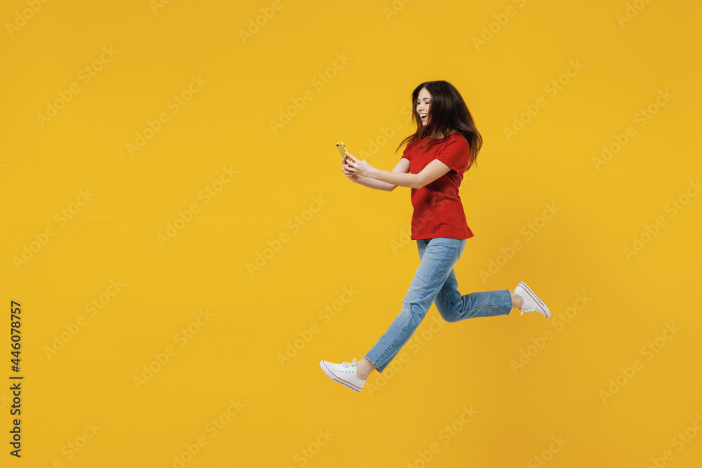Full size body length excited young brunette woman 20s wears basic red t-shirt jump hold in hand use mobile cell phone isolated on yellow background studio portrait. People emotions lifestyle concept. Full size body length excited young brunette woman 20s wears basic red t-shirt jump hold in hand use mobile cell phone isolated on yellow background studio portrait. People emotions lifestyle concept.