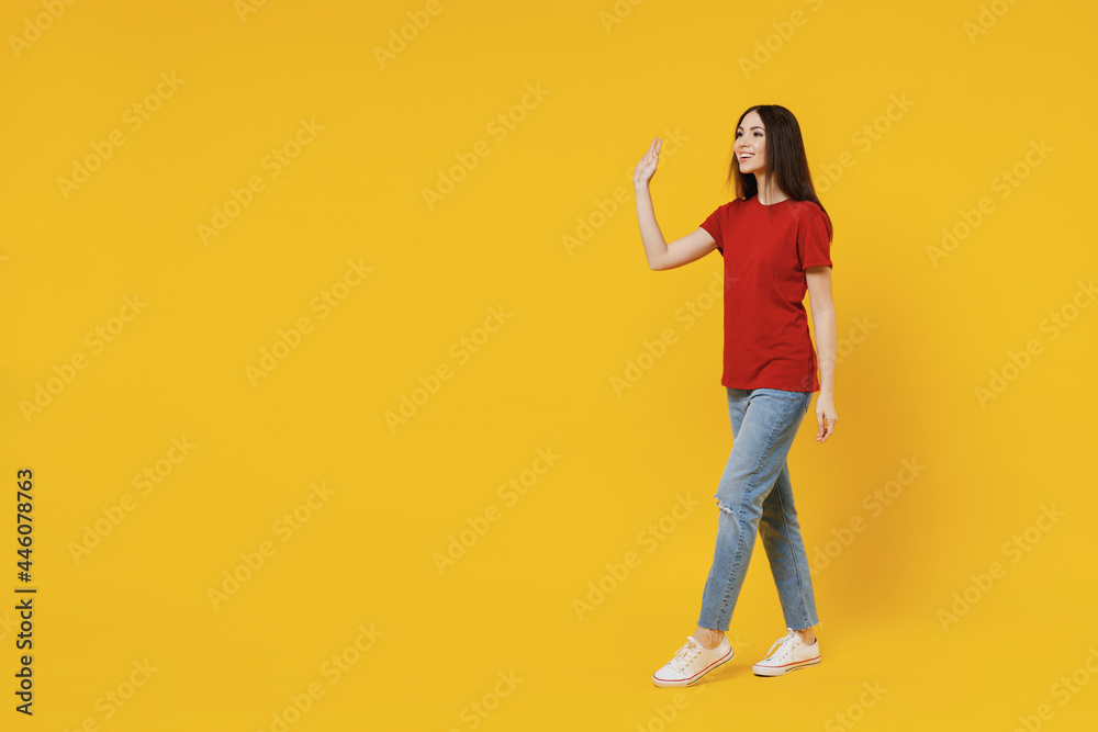 Full size body length side view profile young brunette woman 20s wear basic red t-shirt stand go move step meet greet waving hand isolated on yellow background studio portrait. People emotion concept Full size body length side view profile young brunette woman 20s wear basic red t-shirt stand go move step meet greet waving hand isolated on yellow background studio portrait. People emotion concept