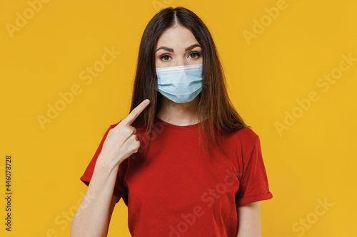 Young brunette woman girl 20s wear basic red t-shirt pointing on sterile face mask ppe to safe from coronavirus virus covid-19 flu on lockdown quarantine isolated on yellow background studio portrait