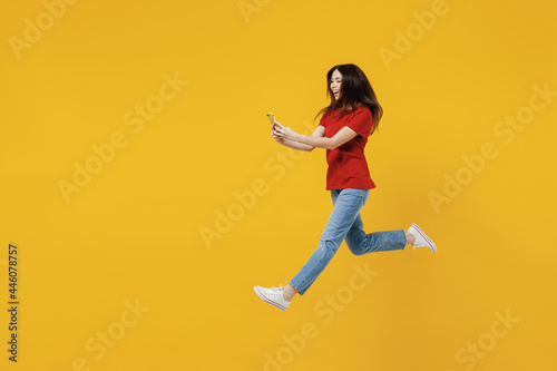 Full size body length excited young brunette woman 20s wears basic red t-shirt jump hold in hand use mobile cell phone isolated on yellow background studio portrait. People emotions lifestyle concept.