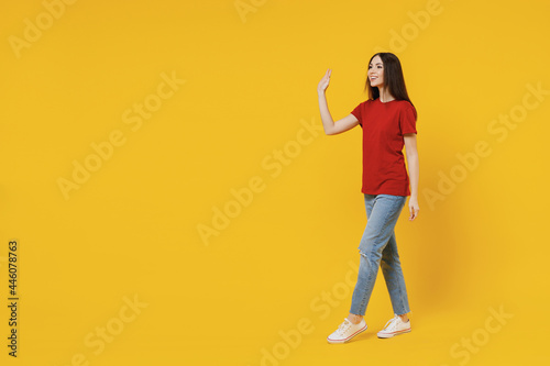 Full size body length side view profile young brunette woman 20s wear basic red t-shirt stand go move step meet greet waving hand isolated on yellow background studio portrait. People emotion concept © ViDi Studio