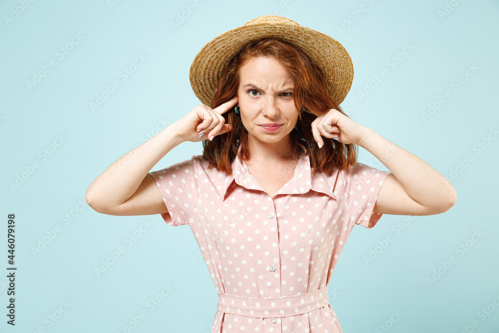 Young redhead curly woman cover ears with fingers do not want to listen wear casual pink dress straw hat look camera isolated on pastel blue color background studio portrait. People emotions concept