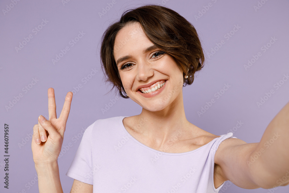 Close up young smiling happy friendly woman 20s with bob haircut wear white t-shirt doing selfie shot on mobile phone show victory v-sign gesture isolated on pastel purple background studio portrait. Close up young smiling happy friendly woman 20s with bob haircut wear white t-shirt doing selfie shot on mobile phone show victory v-sign gesture isolated on pastel purple background studio portrait.