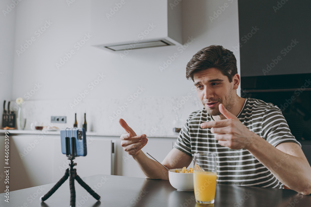 Bottom view young overjoyed man in t-shirt use mobile cell phone watching movie film live stream eat breakfast muesli cereals do winner gesture clench fist sit by table in light kitchen at home alone.