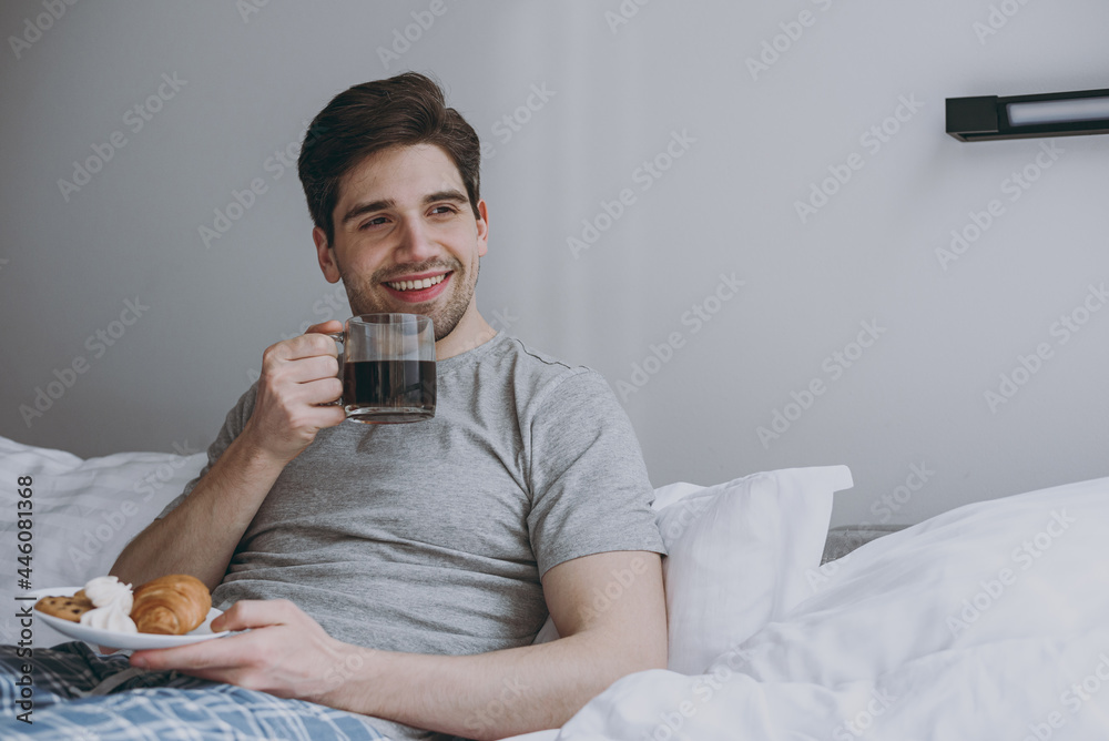 Smiling dreamful minded young man in pajamas grey t-shirt lying sit in bed hold cup drink coffee eat croissant look aside resting relax at home indoors bedroom Good mood night morning bedtime concept Smiling dreamful minded young man in pajamas grey t-shirt lying sit in bed hold cup drink coffee eat croissant look aside resting relax at home indoors bedroom Good mood night morning bedtime concept