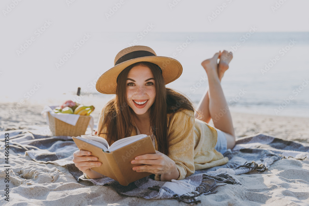 Full body young smiling traveler tourist woman in straw hat shirt summer clothes read book lying on plaid have picnic outdoor on sea sand beach background People vacation lifestyle journey concept