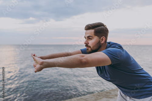 Young strong sporty athletic fit sportsman man in sports clothes warm up training do stretch lunge exercise rising hands up at sunrise sun over sea beach outdoor on pier seaside in summer day morning