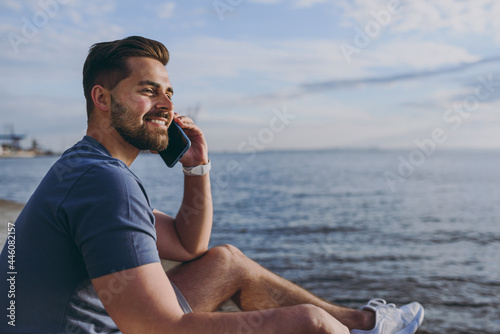 Side view young strong sporty fit fun sportsman man 20s in sports clothes shorts warm up train talk by mobile cell phone at sunrise sun over sea beach outdoor on pier seaside in summer day morning © ViDi Studio