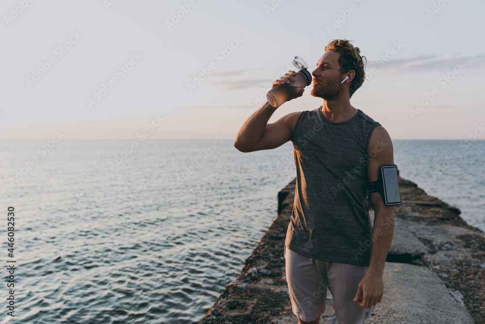 Young strong toned fit sportsman man in sport clothes earphones mobile cell phone in case warm up training drink water at sunrise sun dawn over sea beach outdoor on pier seaside in summer day morning. Young strong toned fit sportsman man in sport clothes earphones mobile cell phone in case warm up training drink water at sunrise sun dawn over sea beach outdoor on pier seaside in summer day morning.