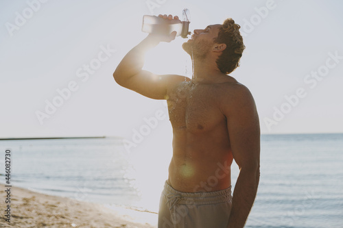 Young thirsty strong sporty toned athletic fit topless sportsman man in sport clothes warm up train drink bottle water at sunrise sun dawn over sea beach outdoor on pier seaside in summer day morning.