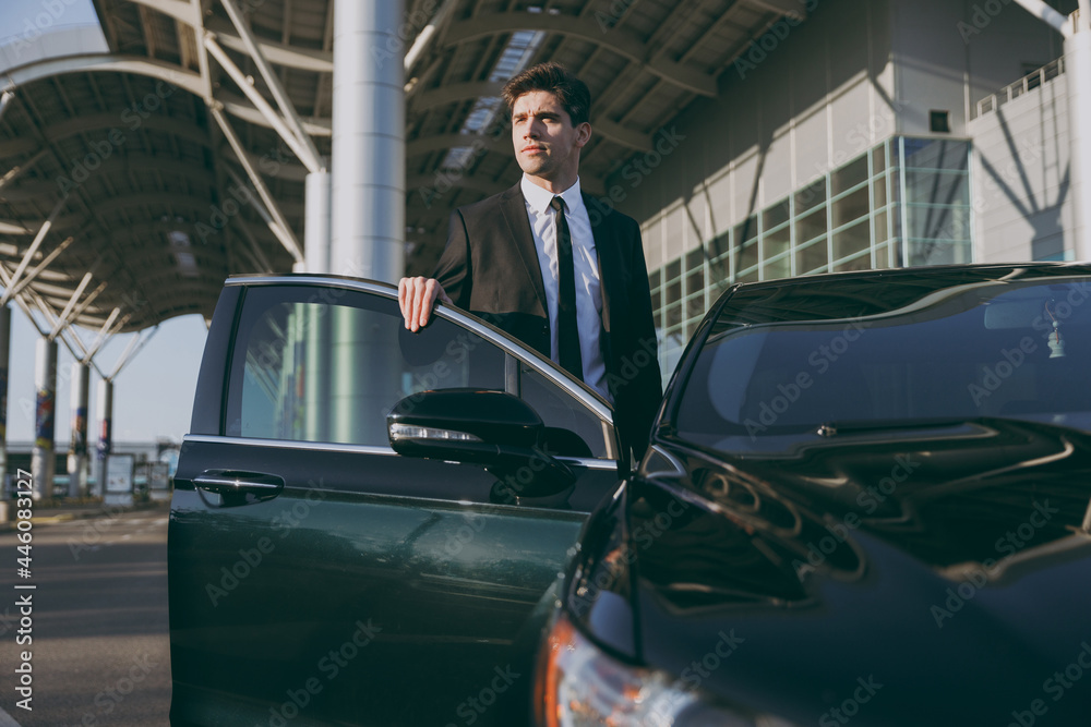 Bottom view serious young traveler brunet businessman man 20s wearing black classic tie suit stand outside at international airport terminal gets into car taxi People air flight business trip concept. Bottom view serious young traveler brunet businessman man 20s wearing black classic tie suit stand outside at international airport terminal gets into car taxi People air flight business trip concept.