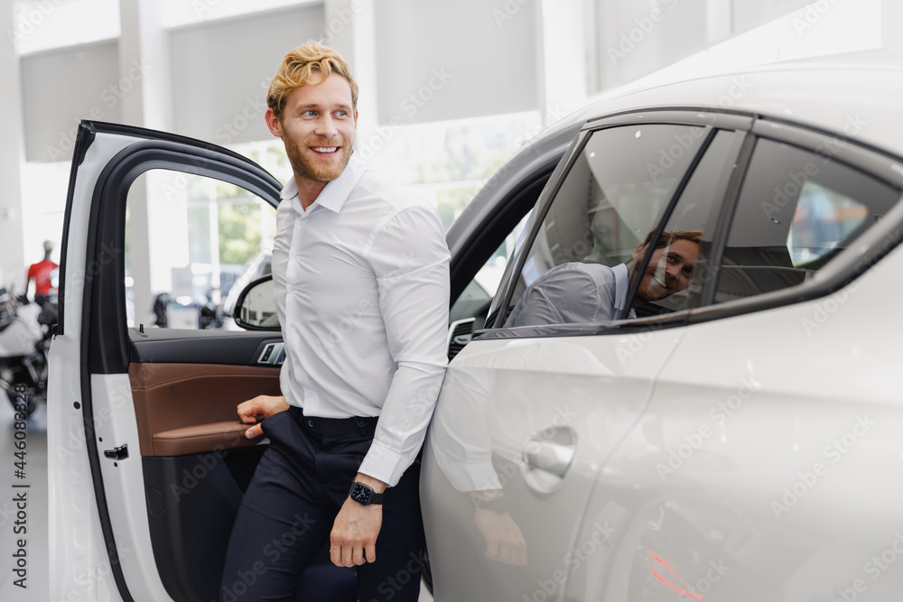 Man smiling satisfied fun customer male buyer client wear white shirt get out car touch door choose auto want buy new automobile in showroom vehicle dealership store motor show indoor Sales concept.