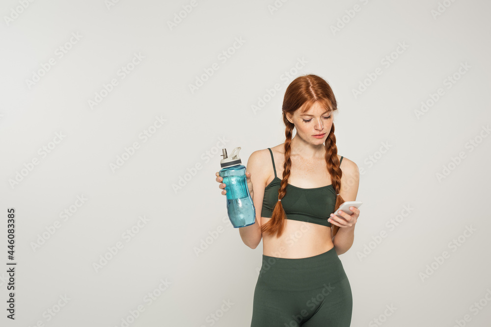 Freckled sportswoman holding sports bottle and using smartphone isolated on grey Freckled sportswoman holding sports bottle and using smartphone isolated on grey
