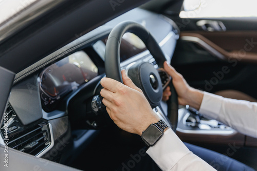 Close up cropped up photo shot hands arms caucasian businessman man wearing white shirt sitting in BMW brand car salon driving hold steering wheel automobile modern vehicle. Car sales driver concept