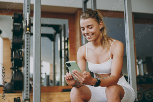 Young happy strong sporty athletic sportswoman woman 20s in white sportswear warm up training sitting near treadmill trainers using mobile cell phone in gym indoors. Workout sport motivation concept