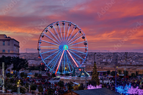 Colorful Ferris wheel against sunset pink sky and Malta cityscape. Christmas market in Valletta Malta aerial view, motion blur, selective focus