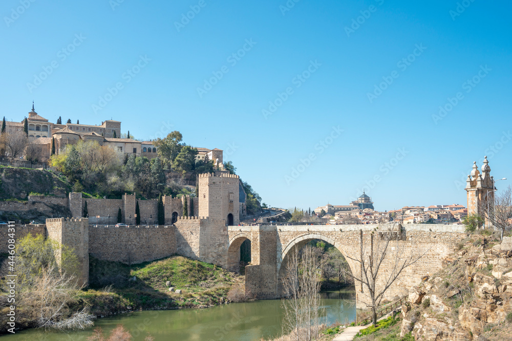 Old bridge of San Martín and in the background the monumental city of Toledo. Beneath the arches of the bridge the river Tagus passes placidly. Old bridge of San Martín and in the background the monumental city of Toledo. Beneath the arches of the bridge the river Tagus passes placidly.
