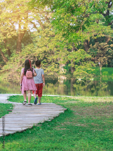 Back view of Asian young girl and boy walking together on pathway through green garden. Sister and brother walking together in park. Happy family spending time together outside in green nature.