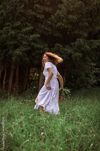 A beautiful happy girl in a long white dress bared her leg, laughing, holding an armful of wild flowers in her hand. Live photo. Selective focus