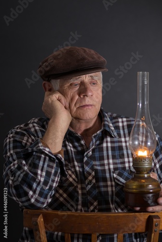 Low key portrait of Ukrainian senior man taking an old kerosene lamp in darkness and thinking while sitting on a wooden chair