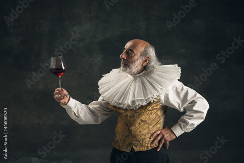 Elderly gray-haired man, medieval hystorical person, actor drinking wine isolated on dark vintage background. Retro style, comparison of eras concept. photo