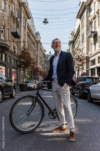 happy middle aged man in blazer and glasses standing with hand in pocket near bicycle on modern urban street
