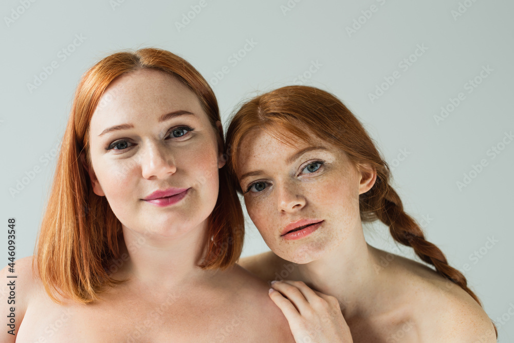 Young freckled woman hugging plus size friend isolated on grey Young freckled woman hugging plus size friend isolated on grey