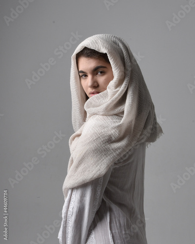 Fototapeta Full length portrait of young woman wearing classical white gown and a head covering veil in biblical style, sitting pose on light studio background