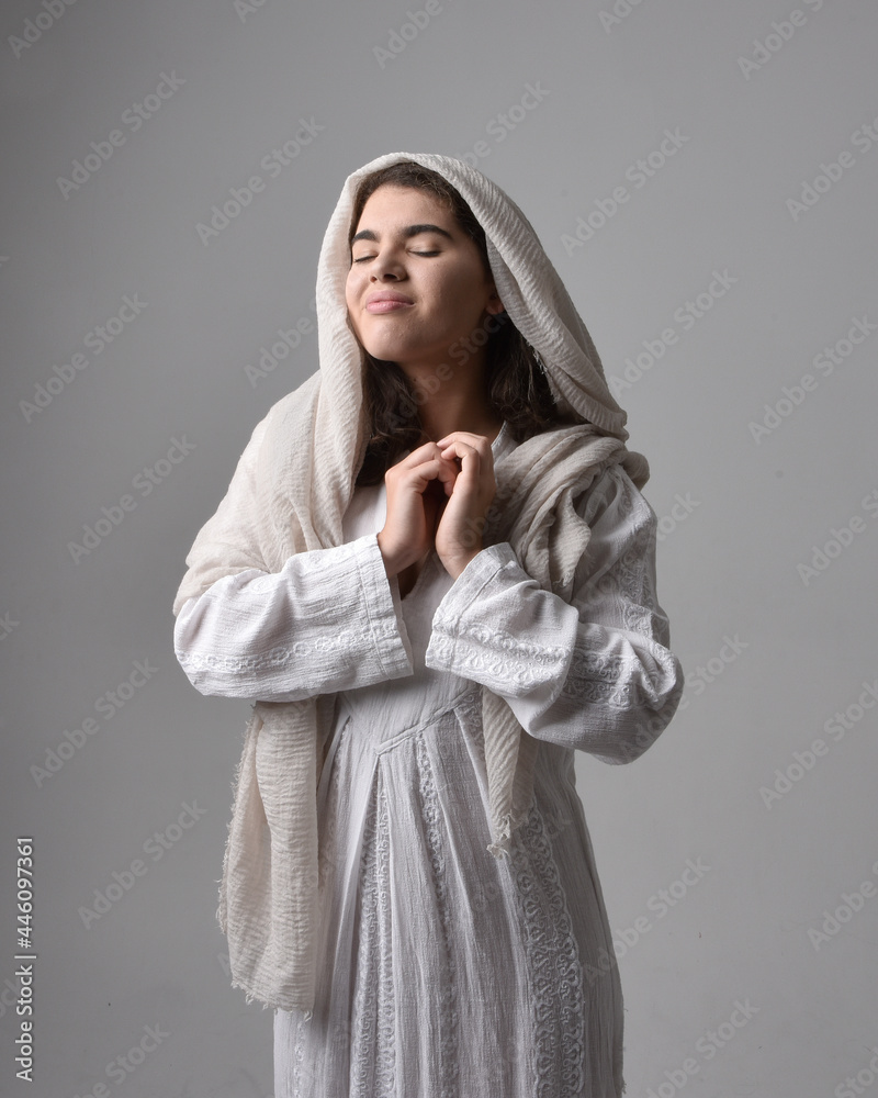 Full length portrait of young woman wearing classical white gown and a head covering veil in biblical style, sitting pose on light studio background. Full length portrait of young woman wearing classical white gown and a head covering veil in biblical style, sitting pose on light studio background.