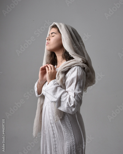 Obraz na plátne Full length portrait of young woman wearing classical white gown and a head covering veil in biblical style, sitting pose on light studio background