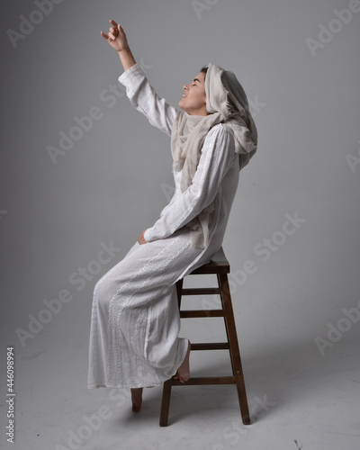 Full length portrait of young woman wearing classical white gown and a head covering veil in biblical style, sitting pose on light studio background.