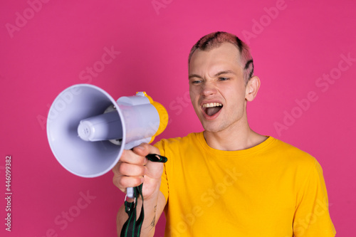 European handsome young man in yellow t shirt on pink background shouting in megaphone