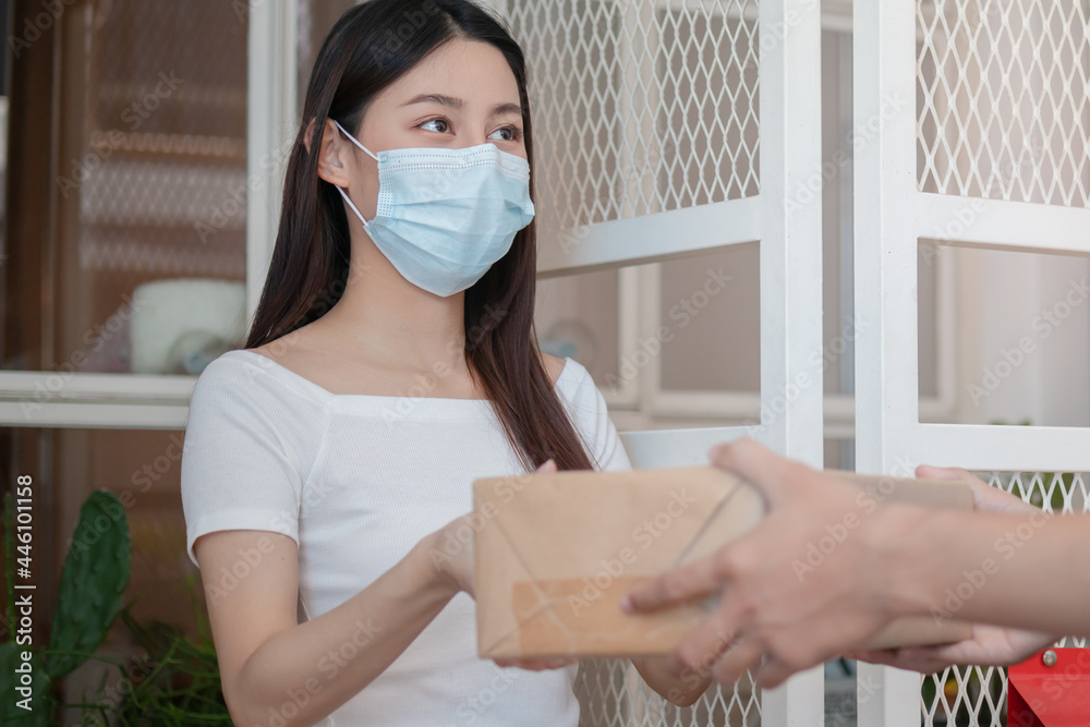 Asain young woman, client received a delivery of cardboard box, parcel from delivery, postman, wearing face mask due to lockdown, quarantine of covid-19 at the door, entrance of her home, house.