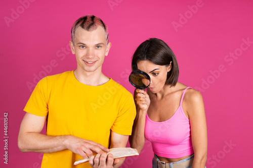 Beautiful modern young couple friends man and woman on pink background holds notebook woman peeks  through magnifier
